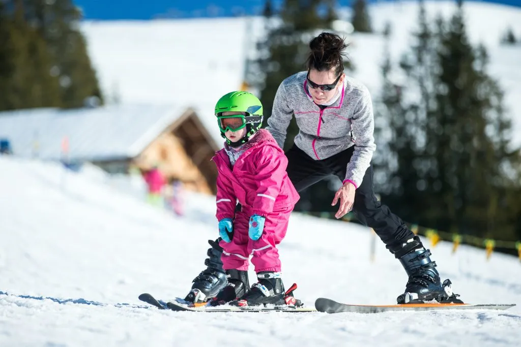Mamma och litet barn åker skidor i Alperna. Aktiv mamma och litet barn med skyddshjälm, skidglasögon och stavar. Skidlektion för små barn. Vintersport för familjen. Liten skidåkare, schweiziska Alperna